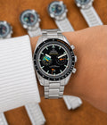 A stainless steel Omega x Swatch Formula 1 wristwatch with a black dial and black bezel, shown on a person's wrist wearing a white knit cuff. The background is a warm brown wood surface with blurred views of other watches.