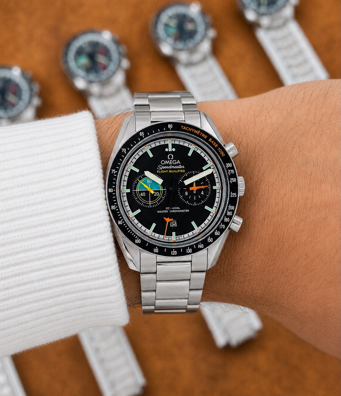 A stainless steel Omega x Swatch Formula 1 wristwatch with a black dial and black bezel, shown on a person's wrist wearing a white knit cuff. The background is a warm brown wood surface with blurred views of other watches.